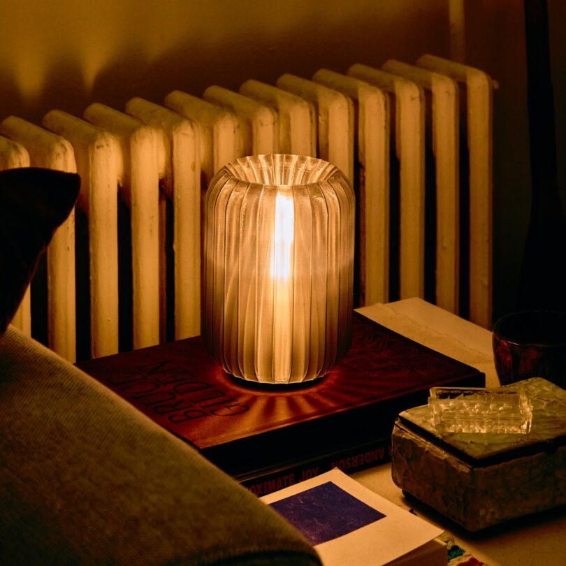 A ribbed glass lamp sits on top of books beside a radiator, casting a warm light in a cozy indoor setting