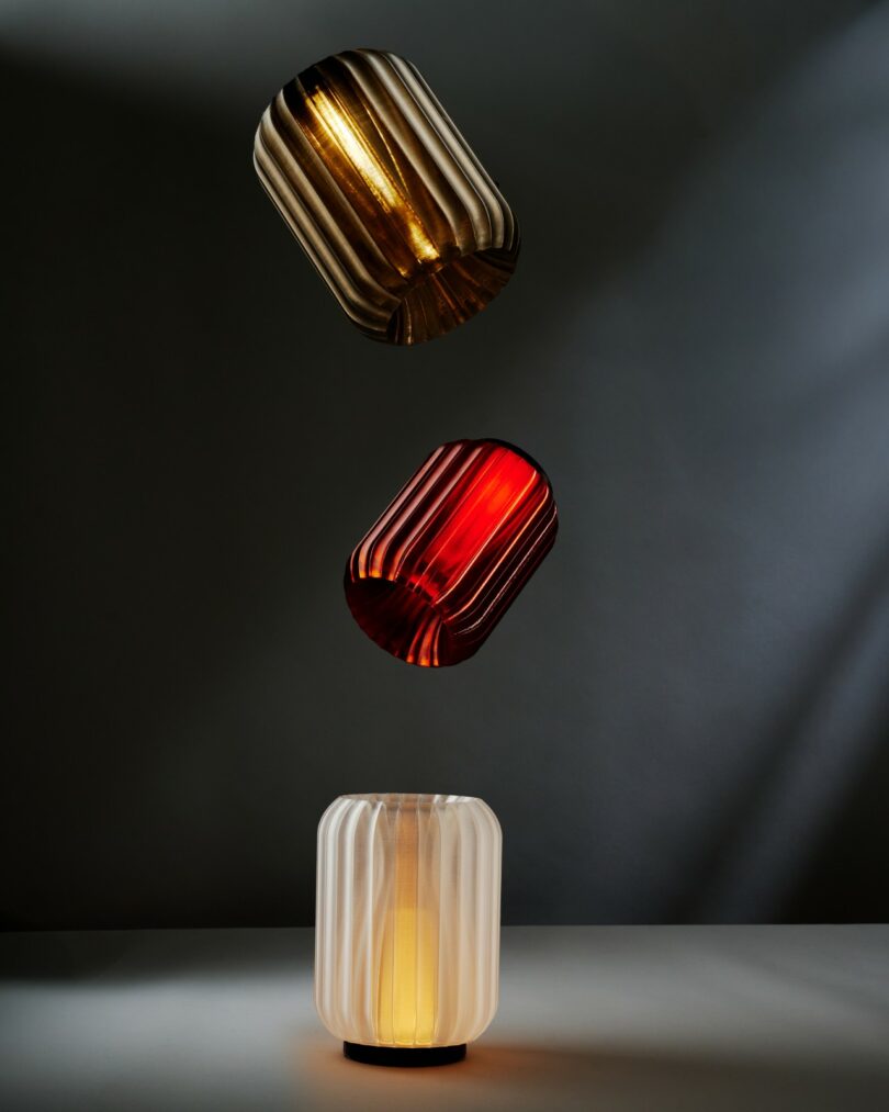 Three ribbed glass lampshades—gold, red, and white—are vertically aligned above a flat surface, each illuminated from within against a gray background