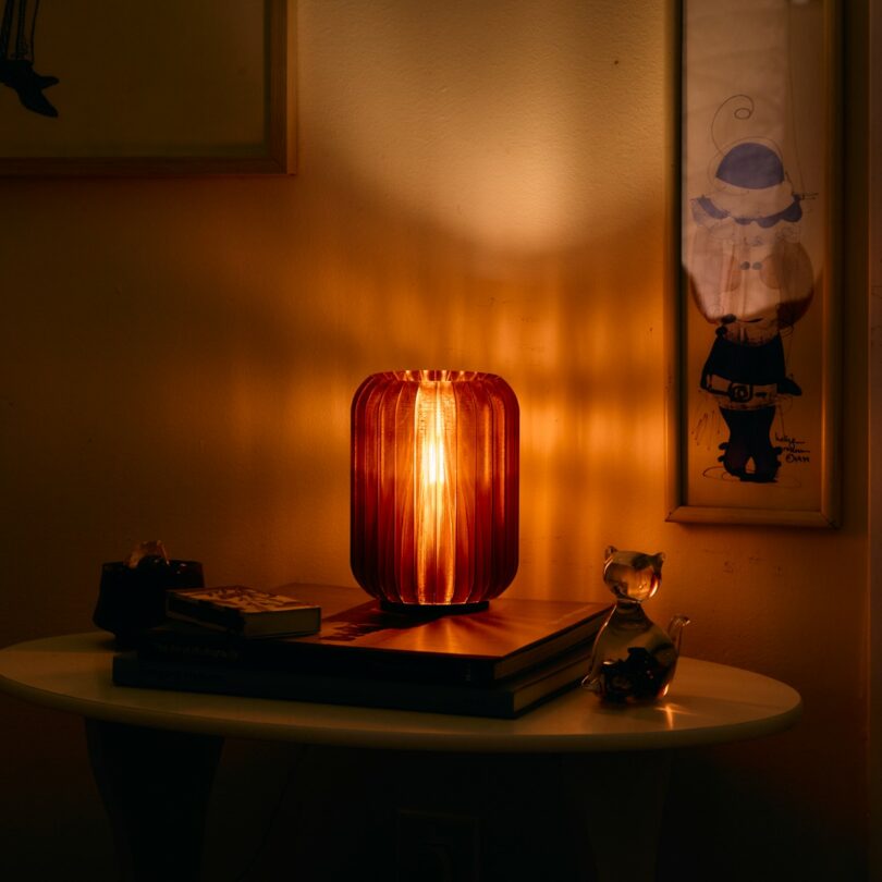 A warm lamp glows on a small table with books, a glass cat figurine, and framed artwork on the wall in a dimly lit room