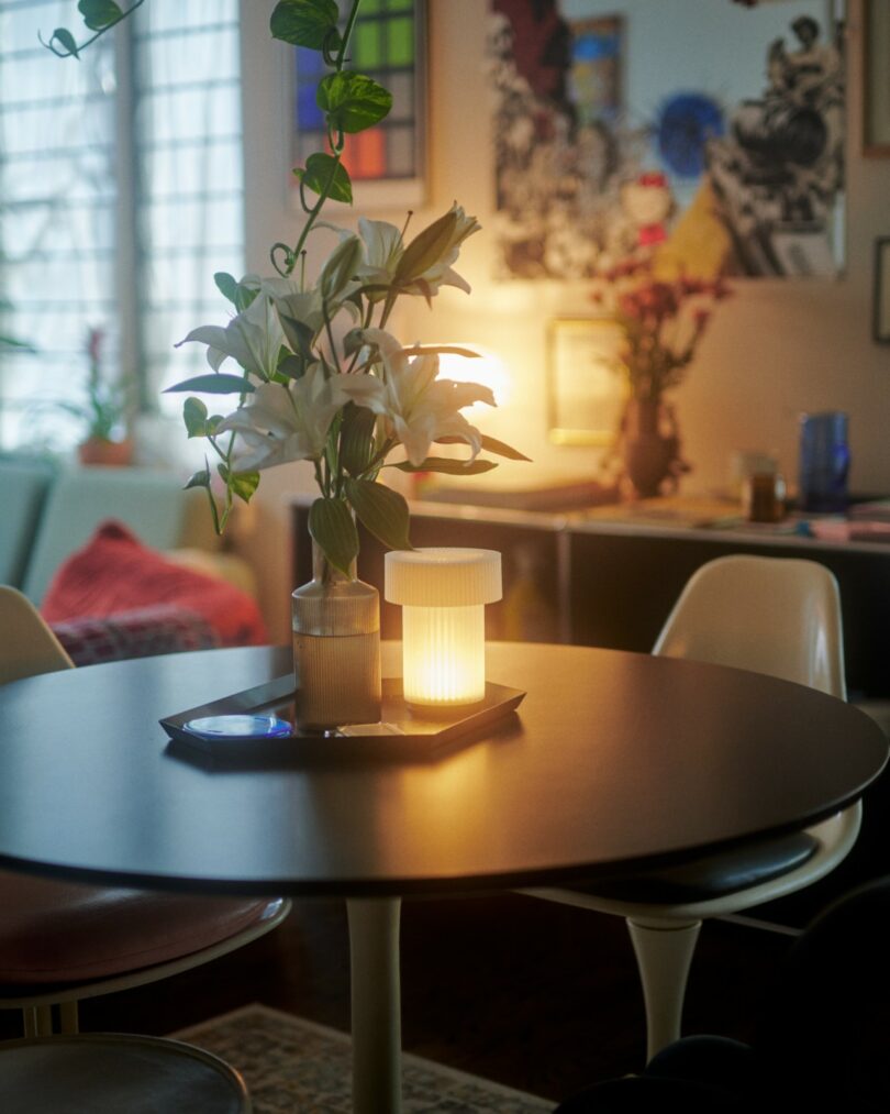 A round black table with a vase of white lilies and a glowing lamp sits in a cozy, softly lit room with modern chairs and framed artwork on the walls