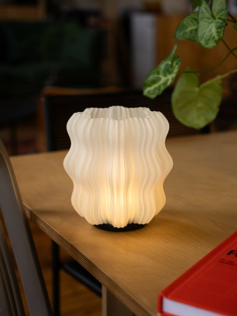 A wavy, white, modern table lamp emits a soft light on a wooden desk, next to a green plant and a red book