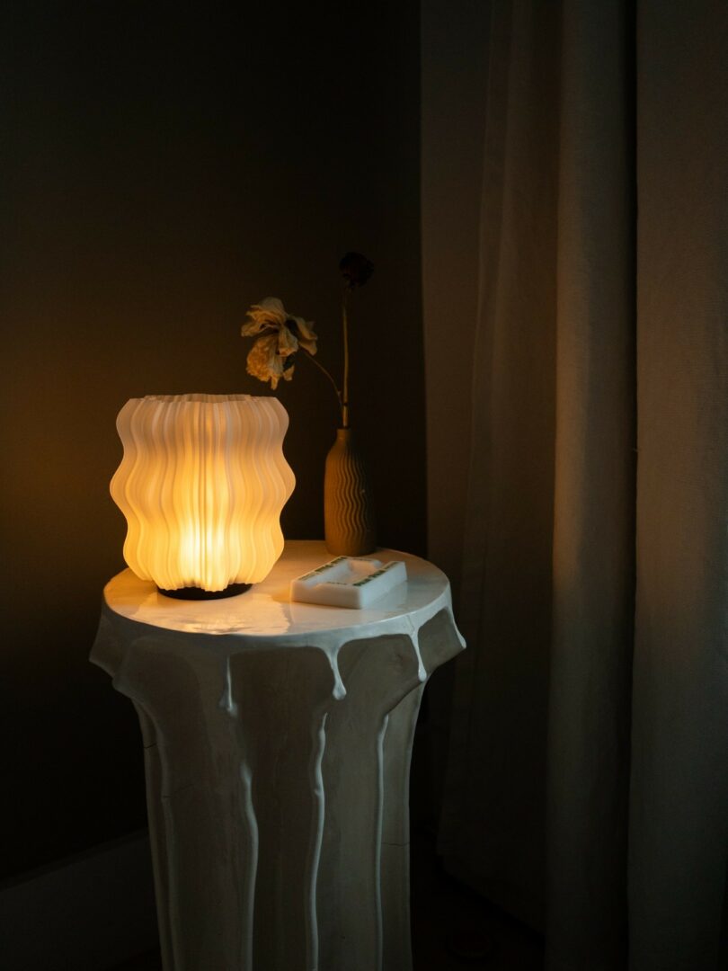A modern wavy lamp emits a warm light on a small round table, next to a vase with dried flowers and a rectangular dish, near a curtain