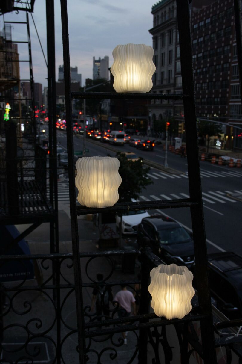 Three wavy, white lanterns hanging on a balcony railing above a city street at dusk, with buildings and traffic in the background