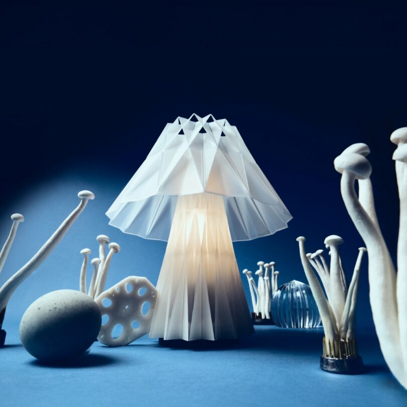 A geometric, origami-style lamp resembling a mushroom is lit amid various artificial mushrooms and abstract objects on a blue surface with a dark blue background