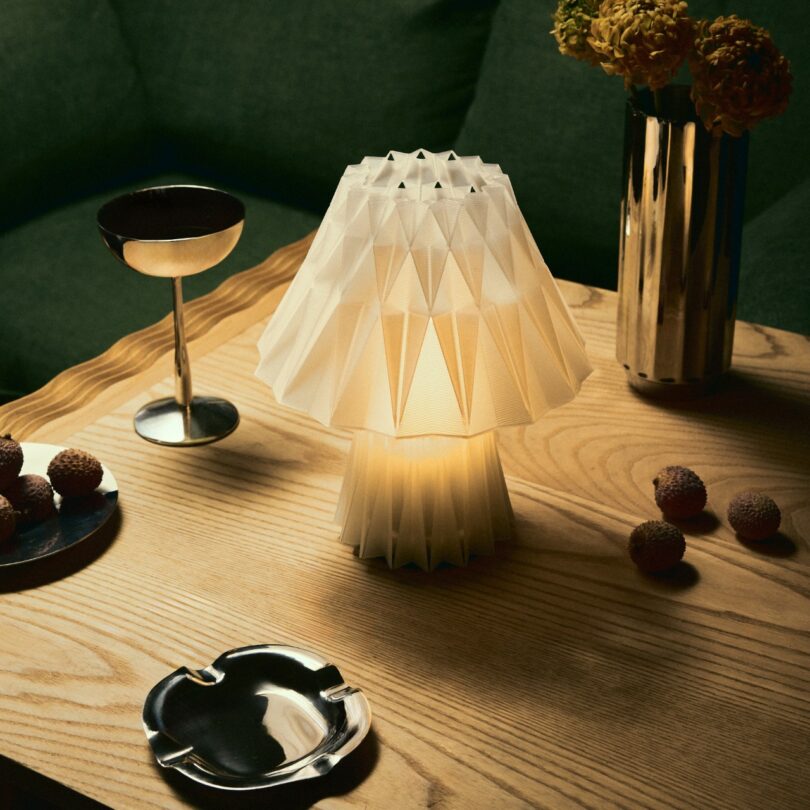 A geometric white table lamp is lit on a wooden table next to a cocktail glass, an ashtray, a vase with yellow flowers, and several round red snacks
