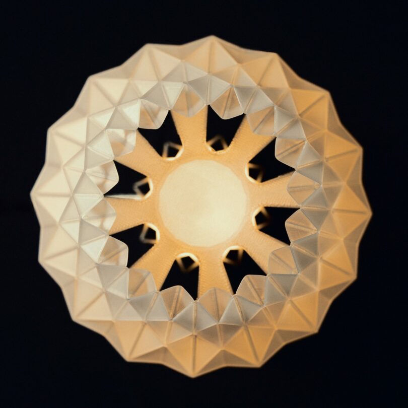 A geometric, patterned lampshade is lit from within, viewed from directly below against a dark background