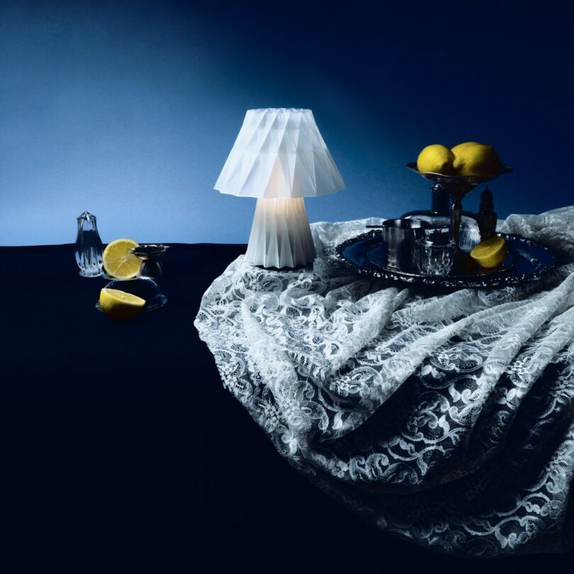 A white table lamp, lace fabric, silver tray with tea set, salt shaker, and lemons are arranged on a dark surface against a blue background