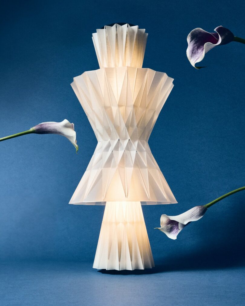 A geometric, white, origami-style lamp glows against a blue background, surrounded by three purple-tipped calla lilies