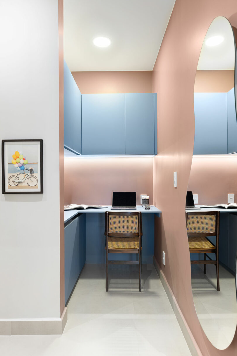 Small modern workspace with blue cabinets, a wooden chair, two laptops on the desk, a wavy wall mirror, and a framed picture of a bicycle with balloons.