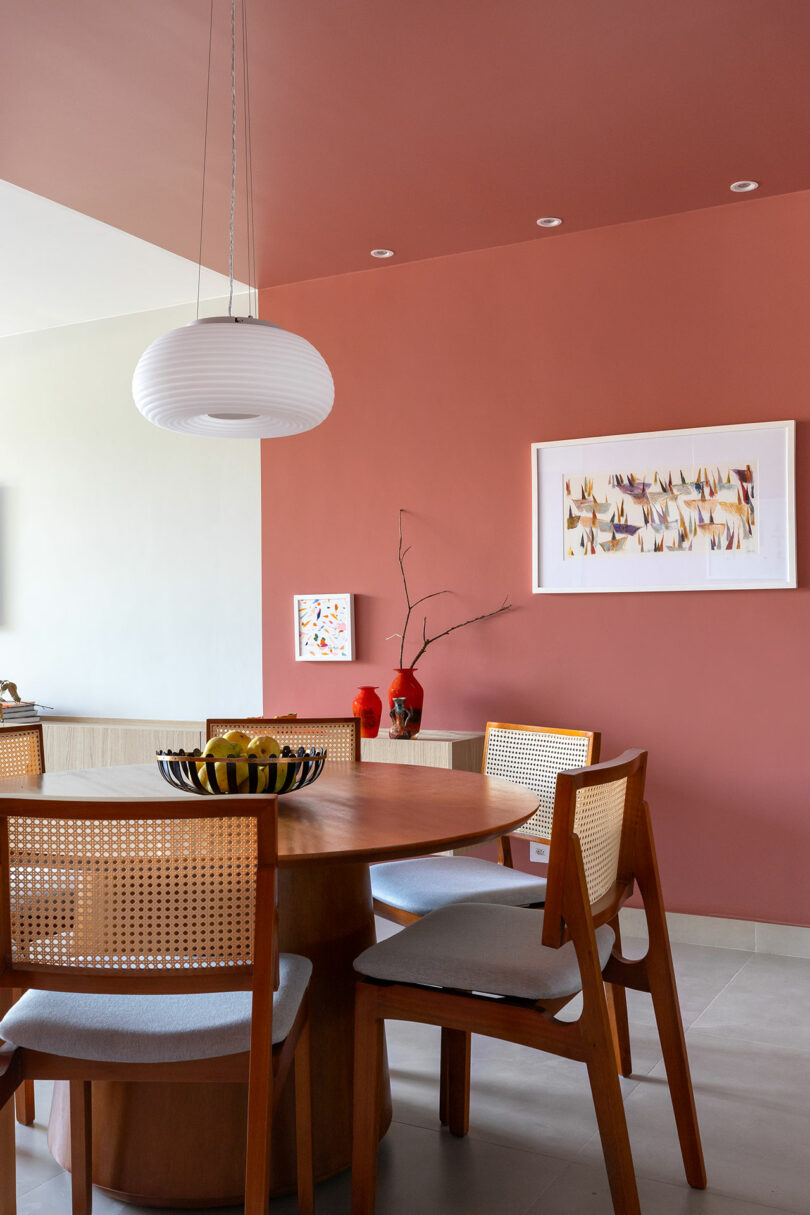 A modern dining area with a round wooden table, four wooden chairs with woven backs, a bowl of fruit, decorative vases, and framed art on a terracotta accent wall.