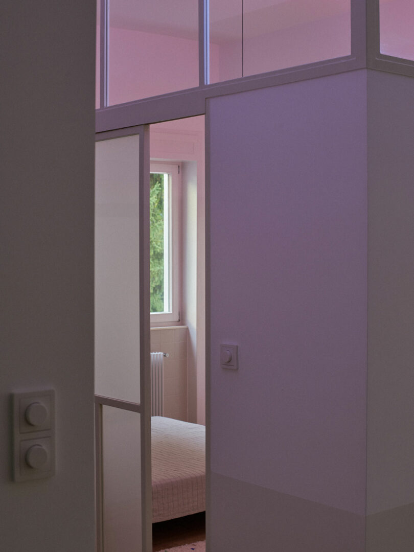A partially open sliding door reveals a bedroom with a window, radiator, and bed, illuminated by soft natural and pink-tinted light.