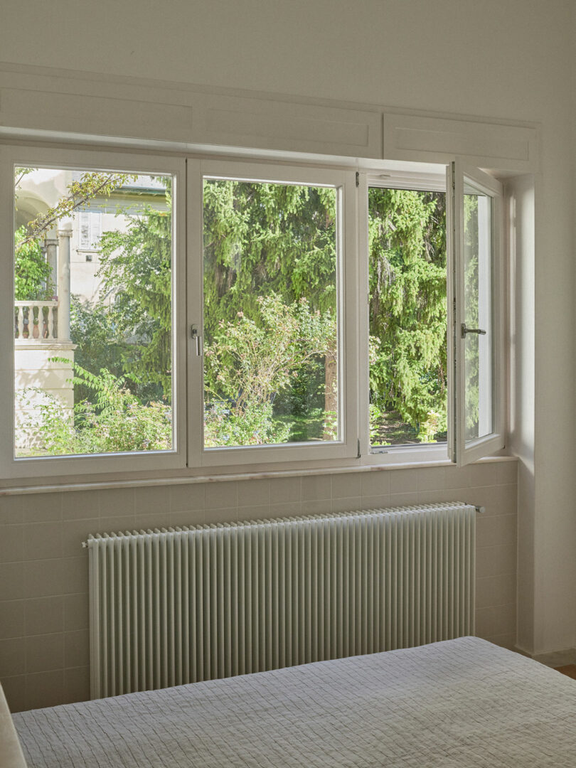 A bedroom with a bed and a large window, partially open, showing green trees and a building outside. A white radiator is below the window.
