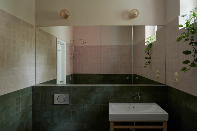 Modern bathroom with green and beige tiles, a white sink, large mirror, wall-mounted toilet, two round lights, and green plants on the windowsill.