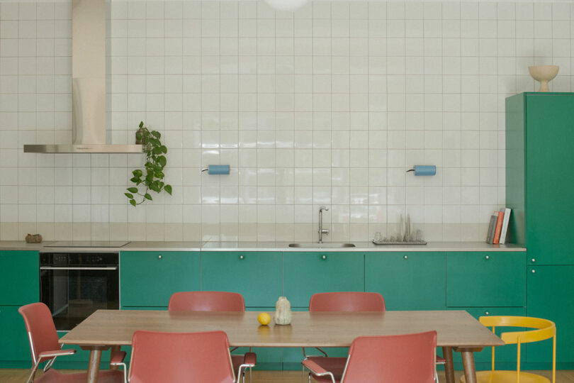 La moderna cocina cuenta con gabinetes verdes, sillas rojas y amarillas, un protector contra salpicaduras de azulejos blancos, una mesa de comedor de madera y una decoración minimalista que incluye libros y plantas en macetas.