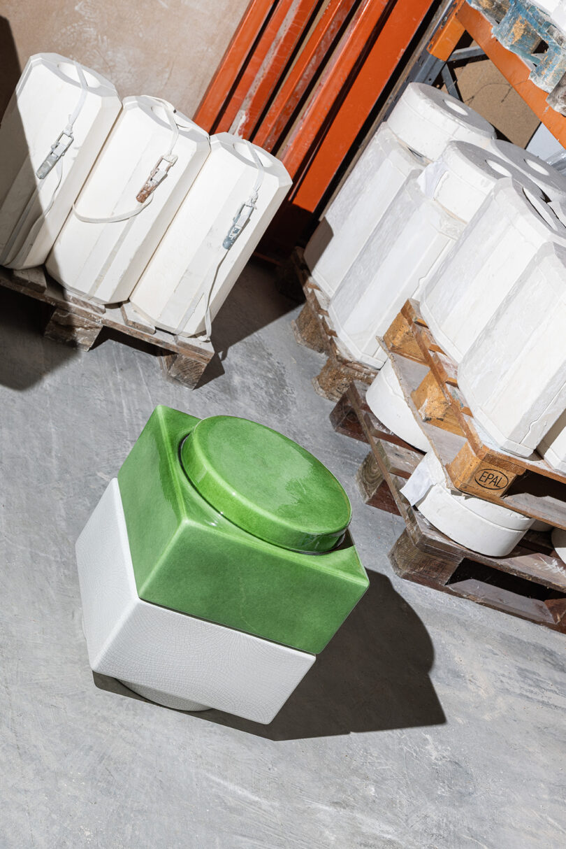 A green and white geometric ceramic object is placed on a concrete floor near white molds stacked on wooden pallets.