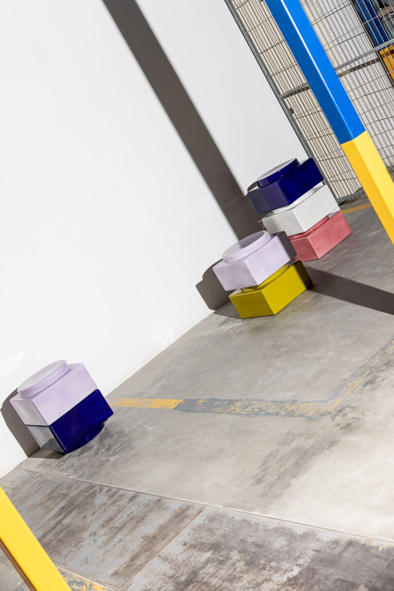 Three colorful, stacked, geometric stools are placed against a wall in a concrete space with yellow and blue metal poles and a wire fence nearby.