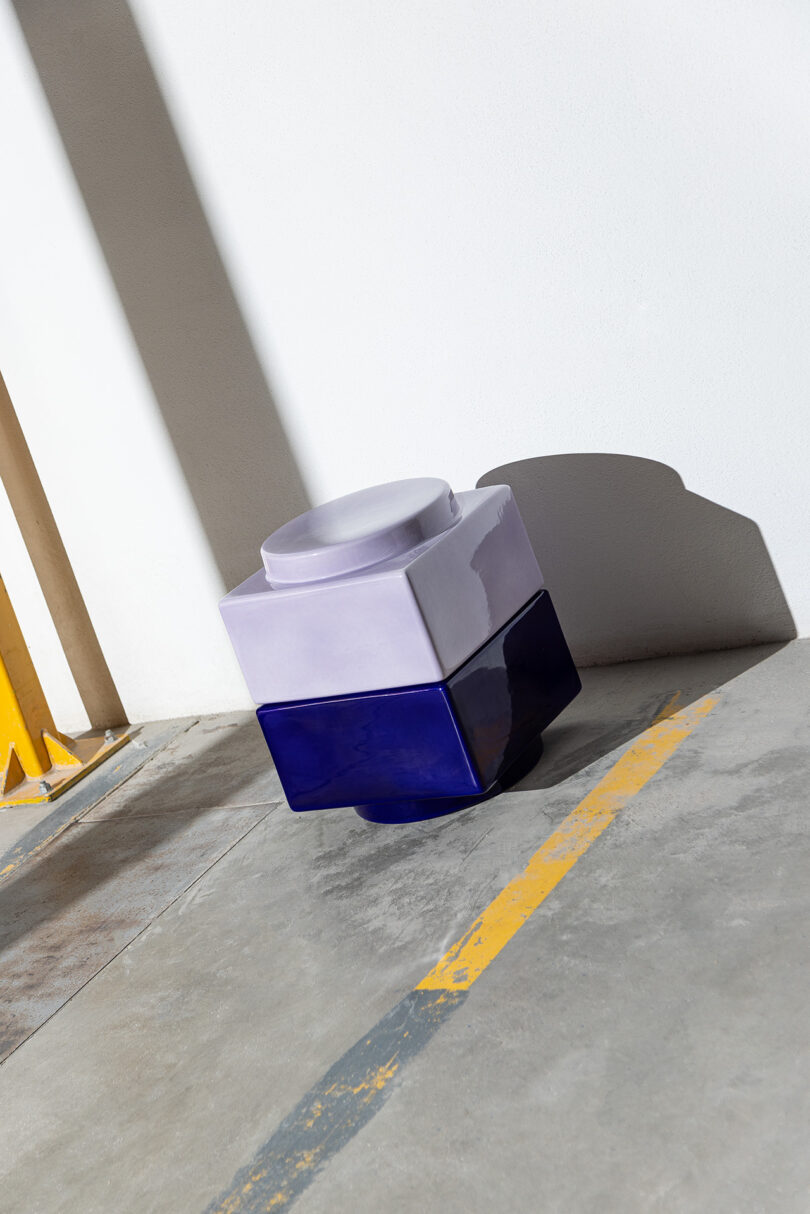 A two-tone ceramic object with a round lid, featuring lavender on top and deep blue on the bottom, sits on a concrete floor near a wall with yellow painted lines.