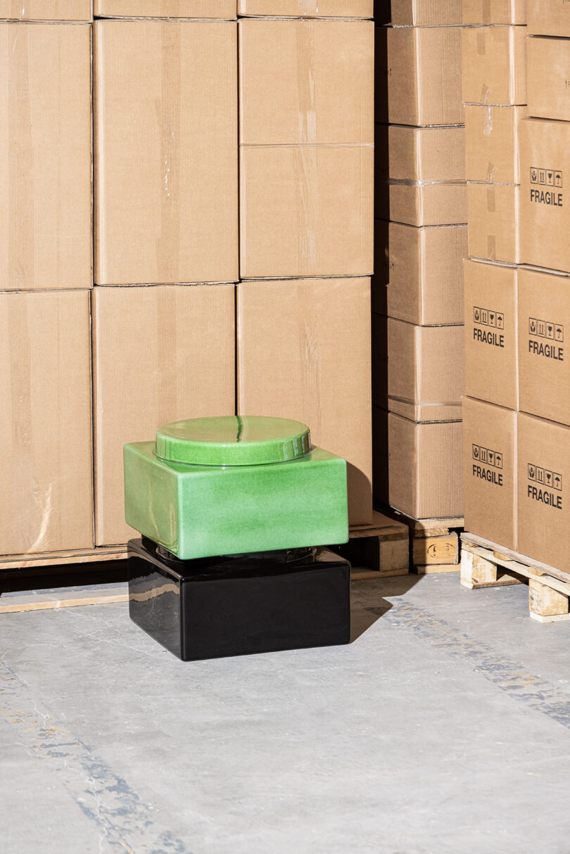 A green, square-shaped ceramic object stacked on a black rectangular base sits on a concrete floor in front of cardboard boxes labeled "FRAGILE" on pallets.