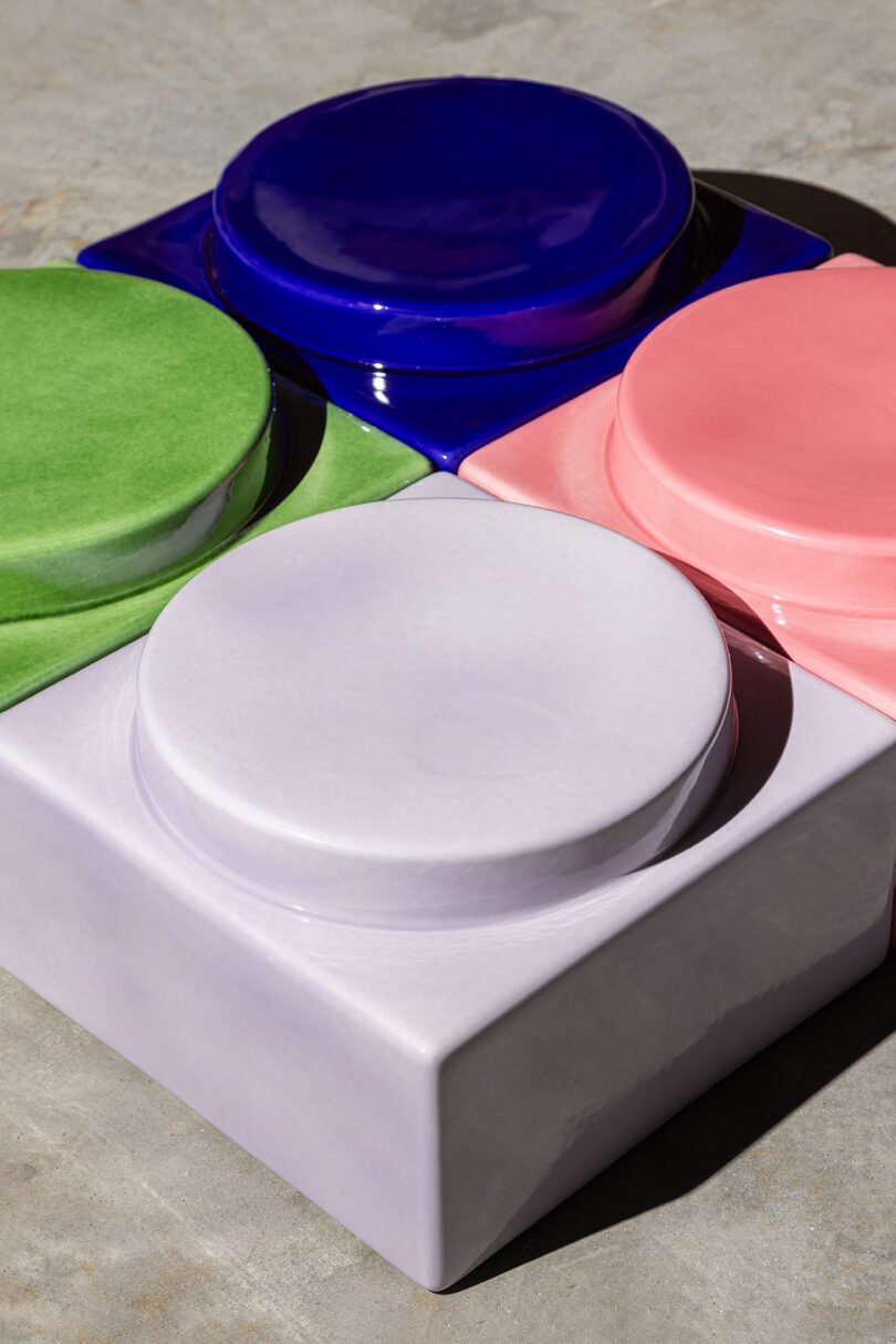 Four glossy, round ceramic shapes in blue, green, pink and lavender are embedded in a square lavender base, set on a concrete surface.