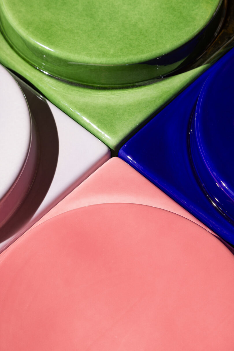 Four ceramic plates in green, blue, white, and pink are arranged closely together, viewed from above, showing their circular shapes and glossy surfaces.