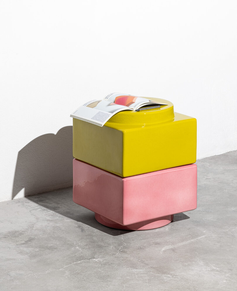 A yellow and a pink geometric ceramic stool stacked on a concrete floor with an open magazine resting on top, against a white wall.