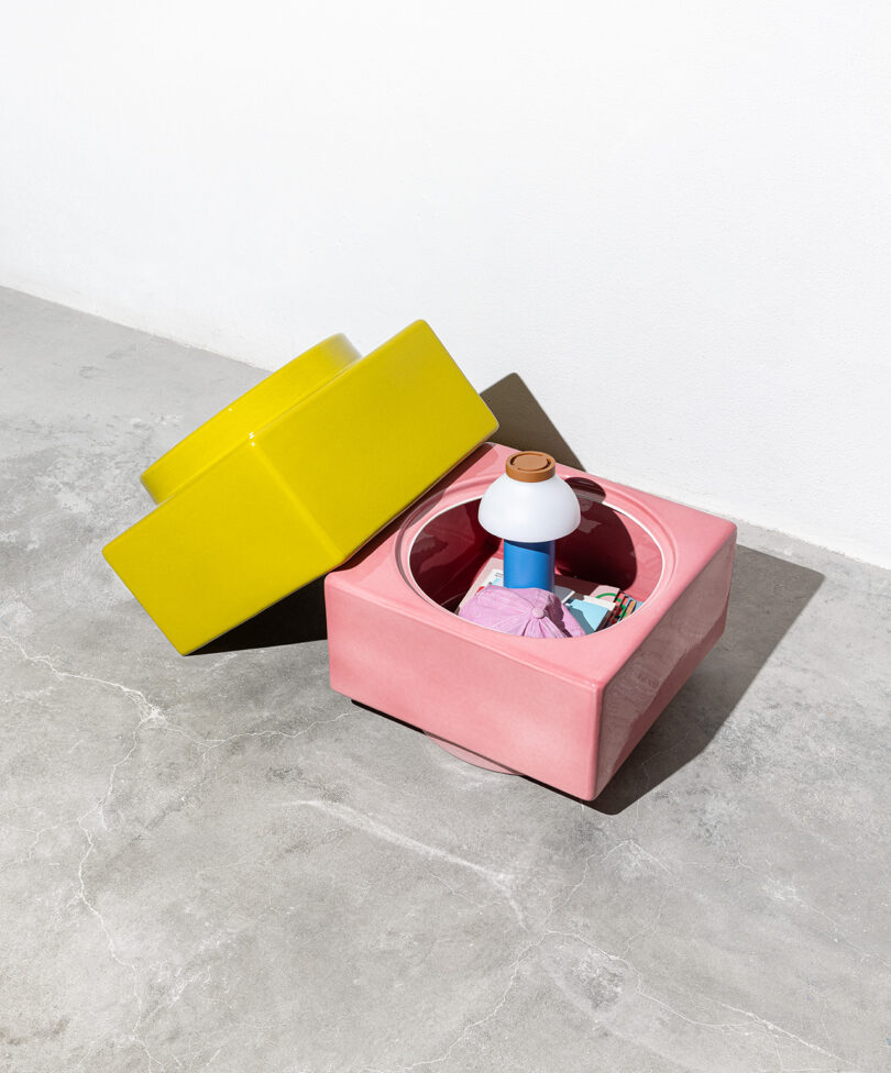 A pink square storage container with its yellow lid beside it, holding various colorful items including a small blue and white lamp, on a gray concrete floor against a white wall.