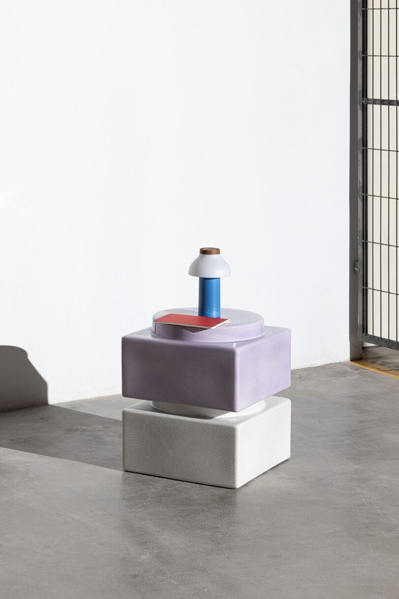 A geometric arrangement of a white square block, a lavender rounded block, and a small blue-and-white vase, displayed on a concrete floor near a wall and metal gate.