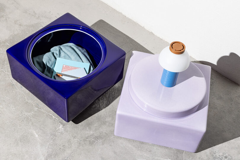 Two square containers on a concrete floor; one blue and open holding clothes and a notepad, the other lavender and closed with a small lamp on top.