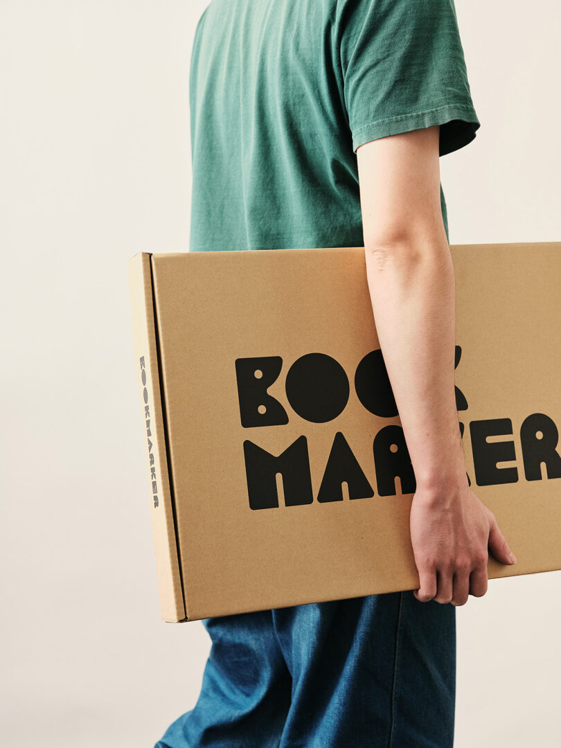 A person in a green shirt and blue pants holds a large rectangular cardboard box labeled "BOOKMARKER" in black letters.
