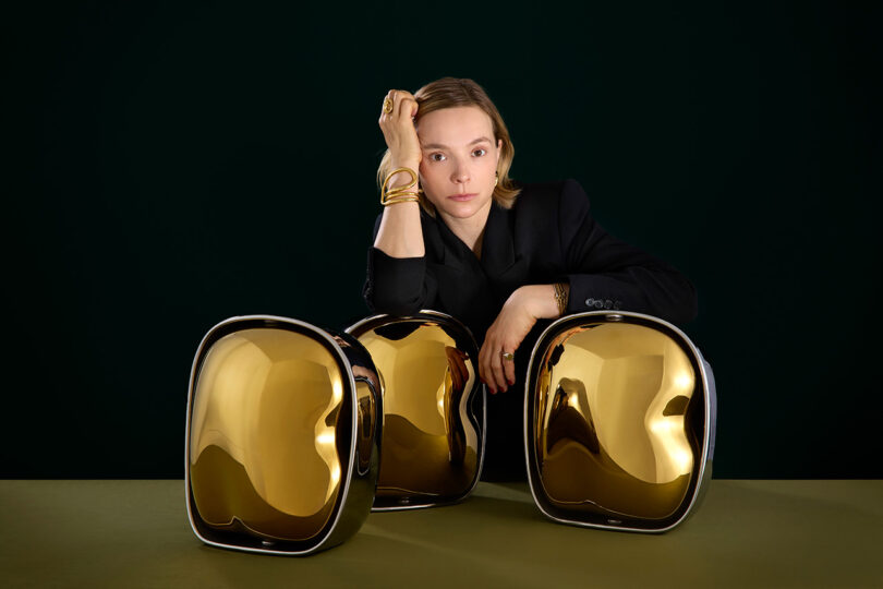 Un hombre con traje negro posa con tres esculturas abstractas doradas brillantes sobre una superficie verde sobre un fondo oscuro.