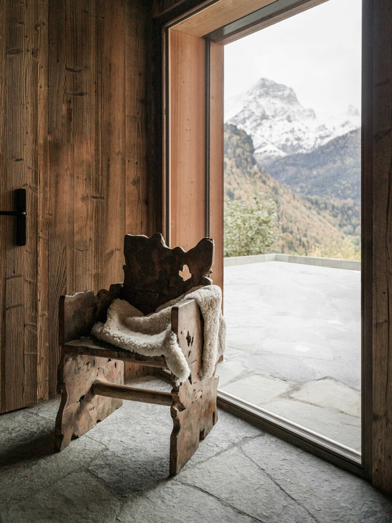 A rustic wooden chair with a sheepskin cover sits next to a large window, overlooking a stone terrace and a view of snow-capped mountains.
