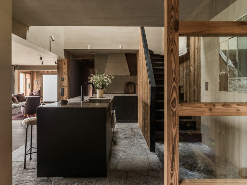 Modern kitchen with dark island, stone floor, wood accents, stairs and glass partition. A vase of flowers sits on the worktop. Living room visible in the background.