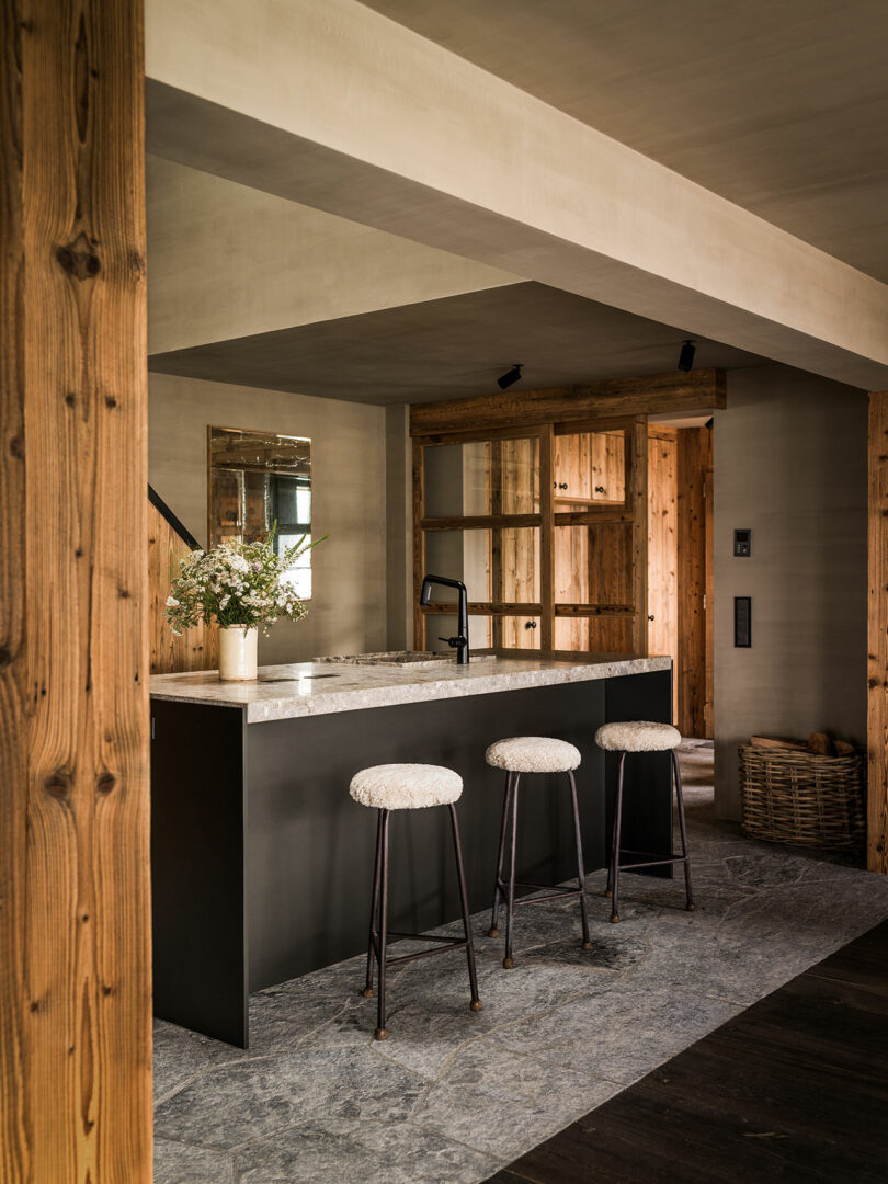 A modern kitchen with a black island, three bar stools with cushions, stone floors, wooden walls, open shelves and a vase of flowers on the counter.