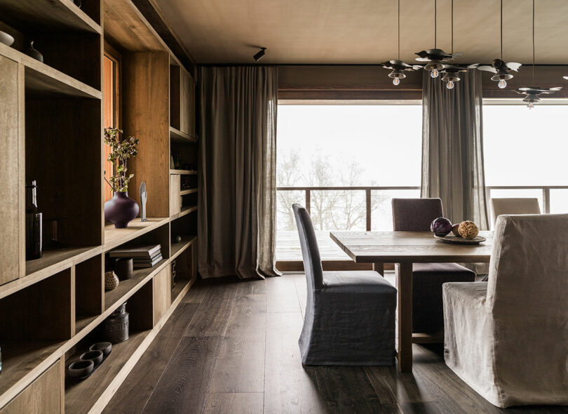 A modern dining room with wooden floors, open shelves, large windows with sheer curtains and a rectangular table surrounded by upholstered chairs.