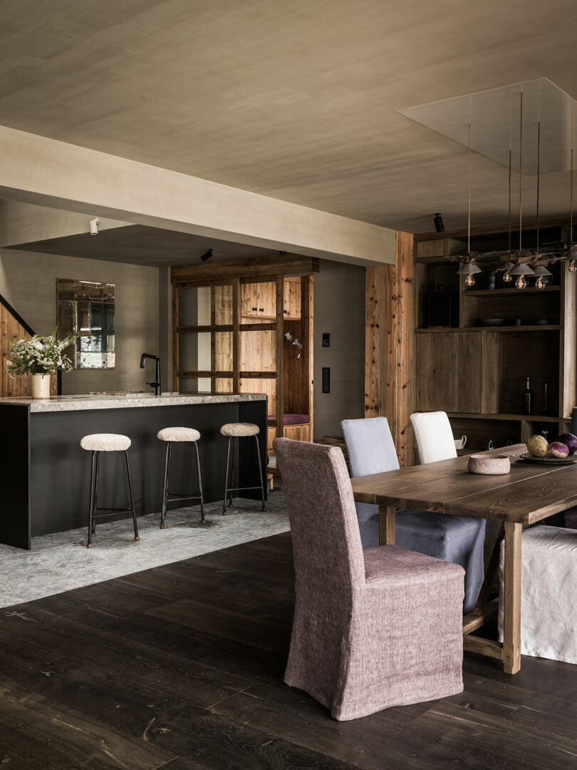 Modern rustic kitchen and dining room with wooden furniture, black island with three bar stools and dining table with upholstered chairs on dark wood floors.