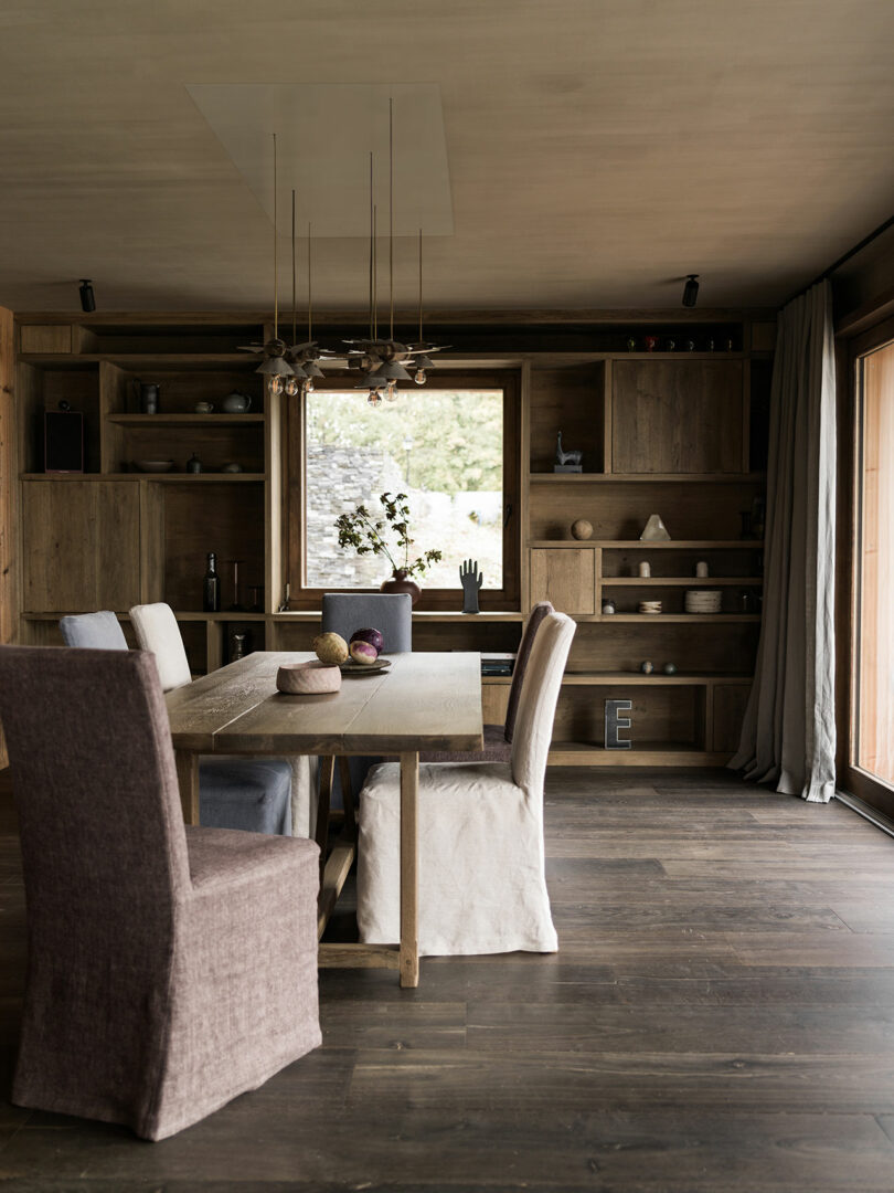 A modern dining room with a wooden table, six fabric-covered chairs, built-in wooden shelves, decorative objects and large windows that let in natural light.