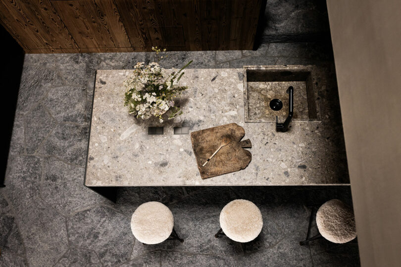 Top view of stone kitchen island with sink, cutting board, knife, flower vase and three round chairs on textured stone floor.