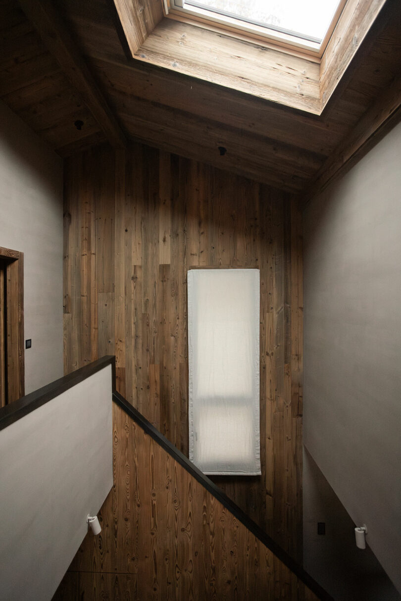A wooden staircase with a large skylight above and a narrow covered window on the wall, with natural wood paneling and minimal decor.