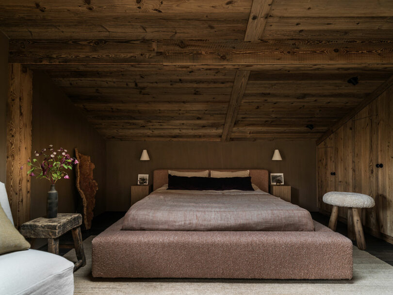 A comfortable bedroom with a large bed, wooden ceiling and walls, two bedside tables with lamps, a stool and a vase of flowers on a rustic side table.