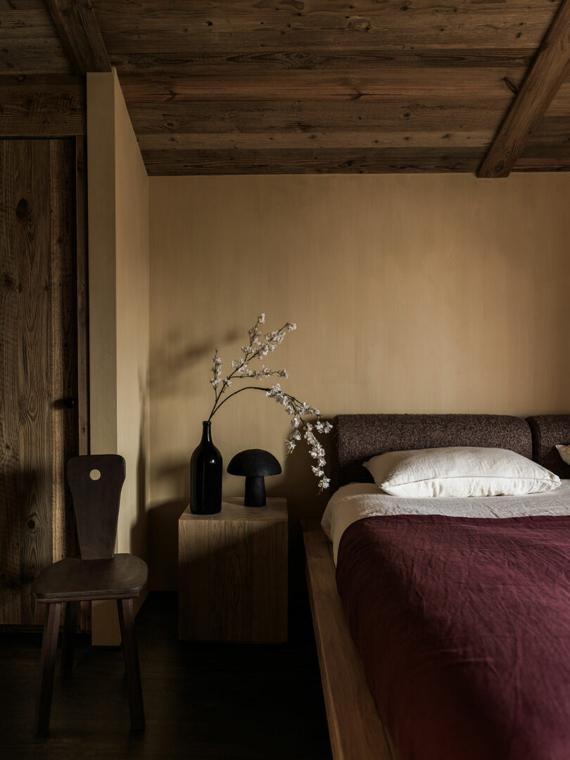 Minimalist bedroom with wooden ceiling, bed with burgundy upholstery, wooden bedside table with black vase with white flowers, black lamp and wooden chair.
