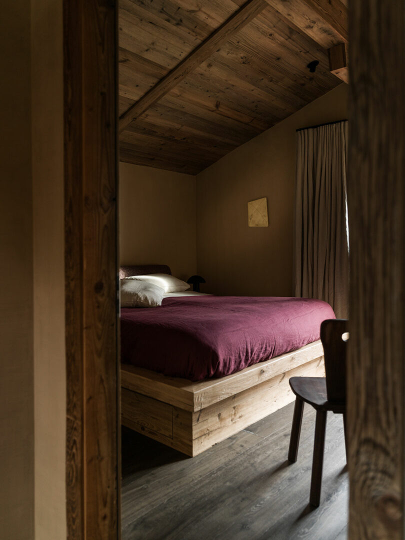 Cozy bedroom with wooden ceiling and floor, bed with burgundy quilt, neutral pillows, wooden chair and beige walls with curtains covering the window.