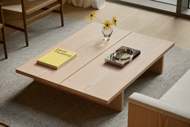 Una mesa de centro de madera clara con un libro amarillo, un jarrón de vidrio con flores amarillas y una bandeja de metal se colocan sobre una alfombra gris en una sala de estar moderna.
