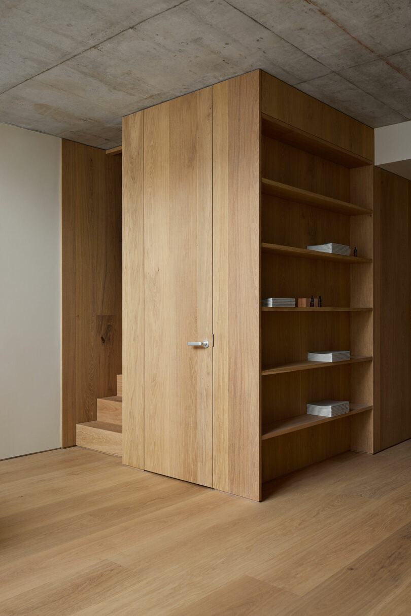 Interior minimalista con paneles de madera, estantes incorporados para guardar algunos objetos pequeños, una puerta cerrada y una escalera parcialmente visible al fondo.
