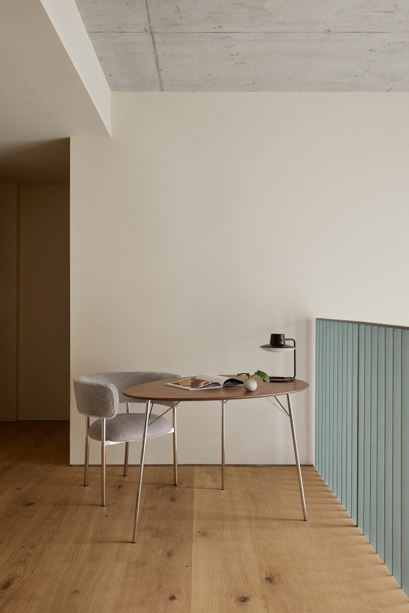 Espacio de trabajo minimalista con mesa redonda, silla gris, libro abierto, planta en maceta y botella de agua negra sobre un suelo de madera clara junto a una barandilla verde claro.