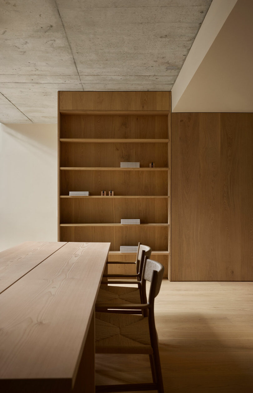 La habitación minimalista está amueblada con una mesa y sillas de madera y una gran estantería de madera con pequeños objetos y libros apoyados en las paredes de colores claros.