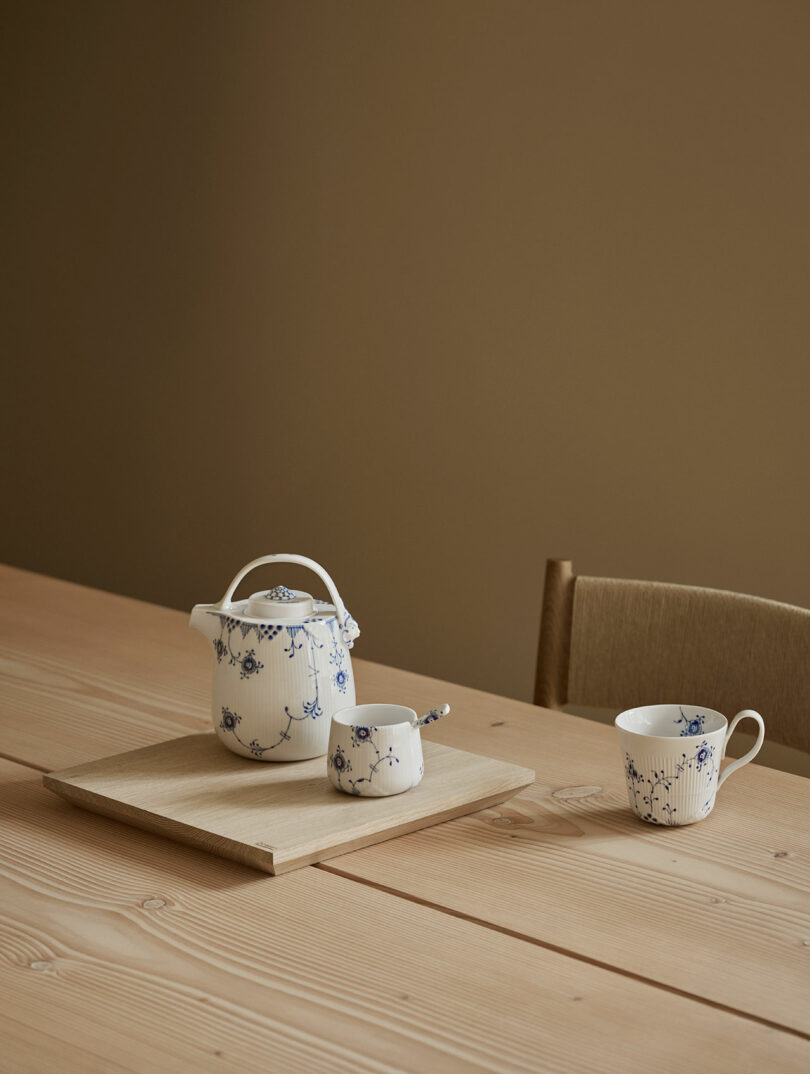 Tetera de cerámica blanca, azucarero y taza de té con estampado floral azul sobre mesa de madera sobre fondo beige.