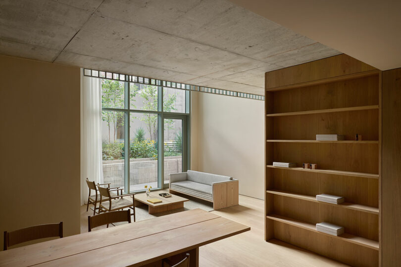 Espacio habitable sencillo con grandes ventanales, muebles de madera clara, estantes empotrados y vistas al pequeño jardín exterior en terraza.