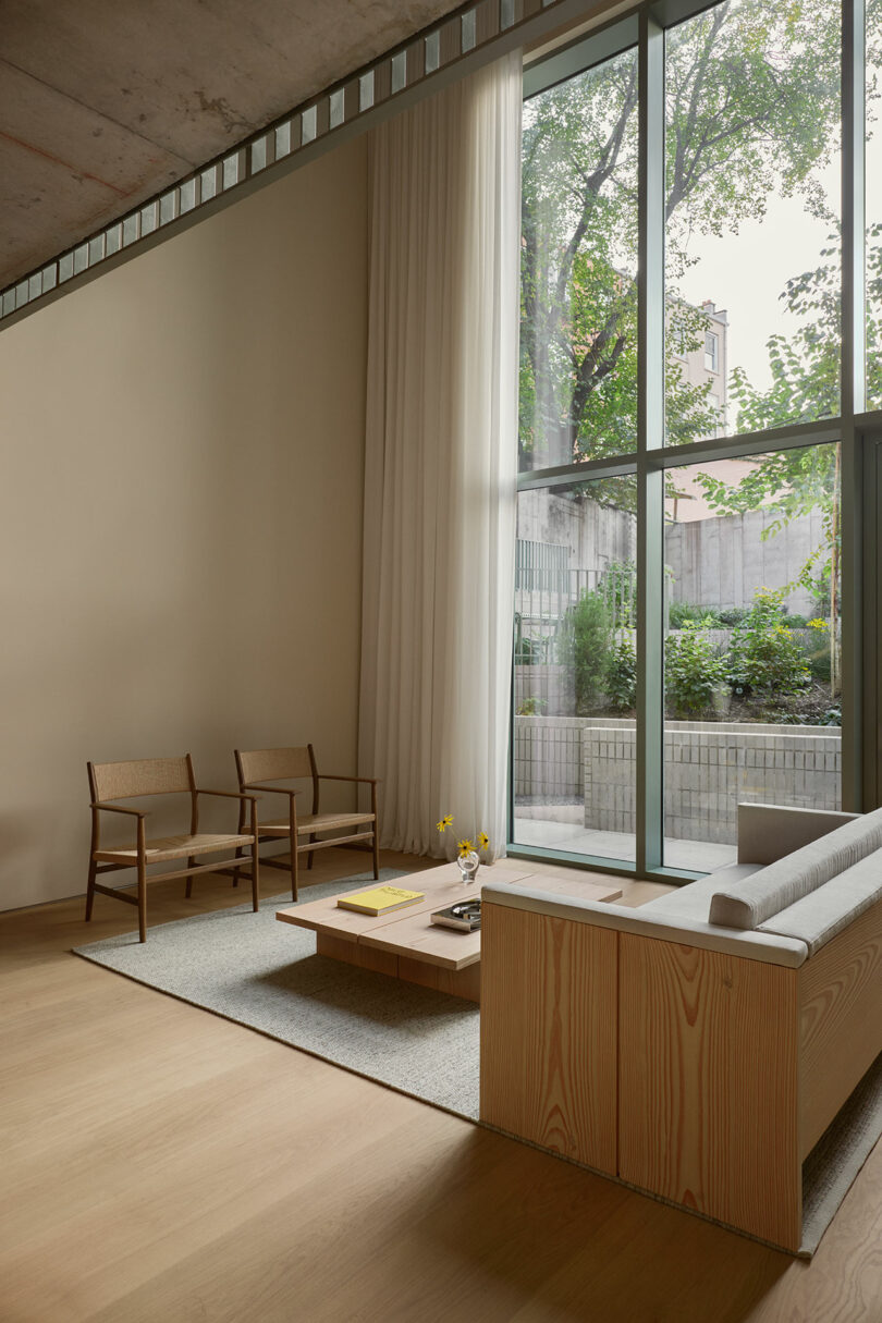 La sencilla sala de estar cuenta con muebles de madera, ventanales, colores neutros y vistas al jardín exterior. La luz natural llena todo el espacio.