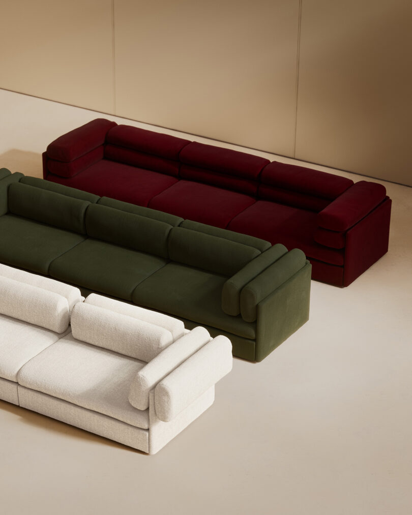 Three modern sofas in white, dark green, and dark red are arranged in a staggered row on a light-colored floor with a beige wall background.