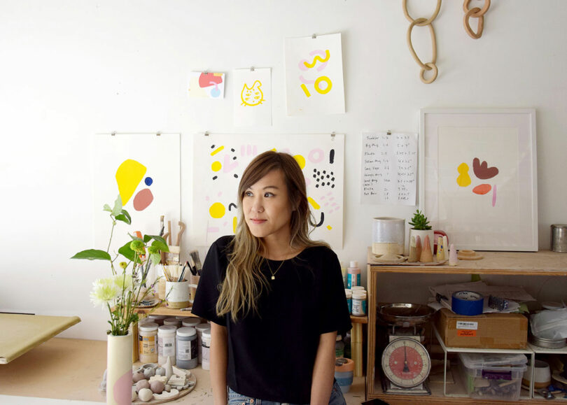 A woman stands in an art studio with colorful abstract artwork on the wall, surrounded by art supplies, plants and ceramics.